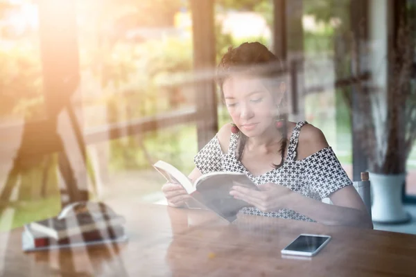 Young woman enjoying reading book sitting near window relaxing in her living with window reflection in the coffee shop