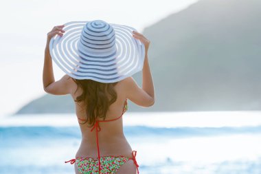 Güneş şapkası ve bikinisiyle sahile bakan bir kadın. Tatil canlılığı sağlıklı yaşam konsepti.