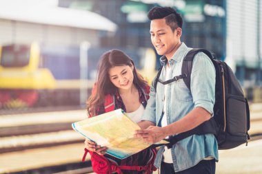 Asyalı genç bir çift haritayı tutuyor ve haritada yeri araştırıyor. Sıcak bir güneş ışığıyla tren istasyonunda iyi yolculuklar. Açık hava macerası tren konseptiyle tek başına seyahat.