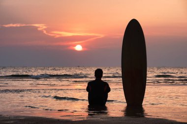 Surfer man sitting alone on the beach looking out to sea with surfboards in the sunset after surfing clipart