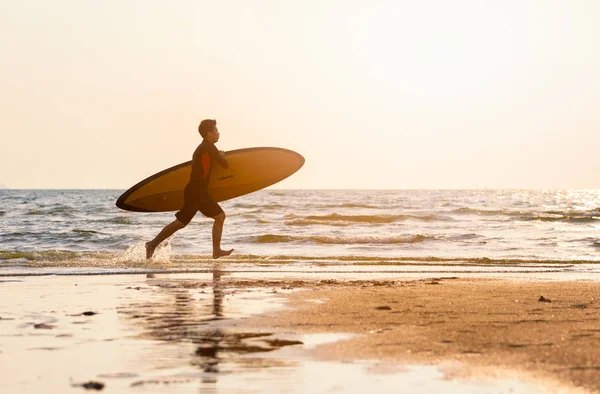 太陽とフレアで海岸を走るサーフボードを持っている若い男 ビーチコンセプトの人々の夏の活動 — ストック写真