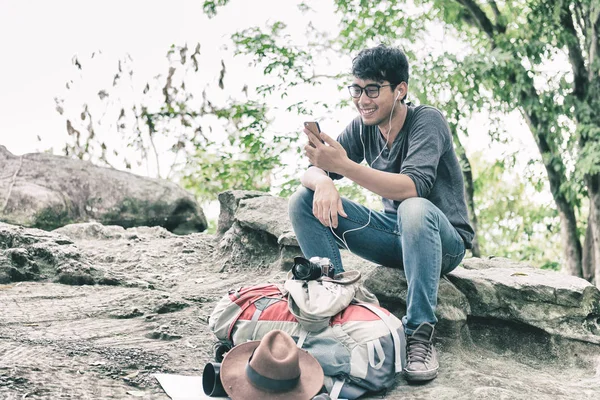 Asian Man Happiness using Smartphone Sitting Alone on Rock in Forest Listening Music Earphones. Having Rest Outdoor. Nature and Modern Technologies and Lifestyle Communications concept