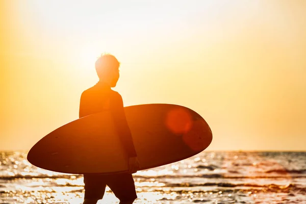 太陽の光とフレアで海岸を歩くサーフボードを持つシルエットの若者 ビーチコンセプトの人々の夏の活動 エクストリームスポーツコンセプト — ストック写真