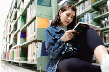 Kütüphanede yerde oturan Asyalı kız öğrenci, Uluslararası Kolej/Üniversite Kütüphanesi'ndeki kitaplığından ders kitabı açma ve öğrenme.
