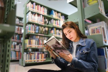 Kütüphanede yerde oturan Asyalı kız öğrenci, Uluslararası Kolej/Üniversite Kütüphanesi'ndeki kitaplığından ders kitabı açma ve öğrenme.