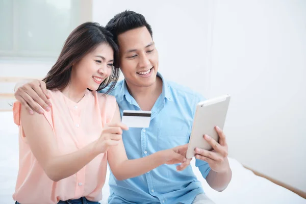 Asian young couple using a tablet for shopping with a credit card while sitting on the bed in the bedroom. High-speed internet 5G service technology at home, Telecom networks designed concept.