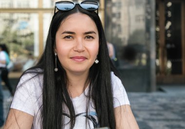 Siyah uzun saçlı çekici mutlu hipster kız oturan portresi. Güzel bir gülümseme ile neşeli esmer kadın bir sokak kafede onu rekreasyon zaman zevk