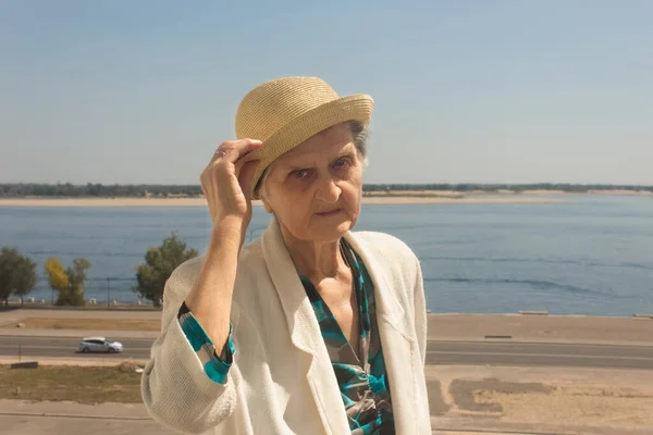 Yaşlı bir kadın yürüyüşe çıktı. Hasır şapkalı, beyaz ceketli, mavi gökyüzü ve nehir altyapısı olan yaşlı bir kadın. Sosyal uzaklık.