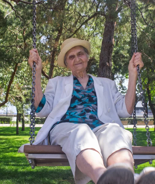 Happy elderly woman has  a fun  on a swing. Senior woman in straw hat and white jacket swings. Social distance.