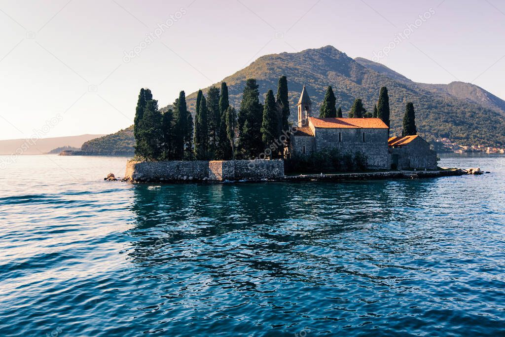 Catholic red roof medieval monastery, bell tower, masonry wall and cypress trees on an island in the Adriatic Sea.  Little island in blue water of Kotor bay among Montenegro mountains. Island panorama