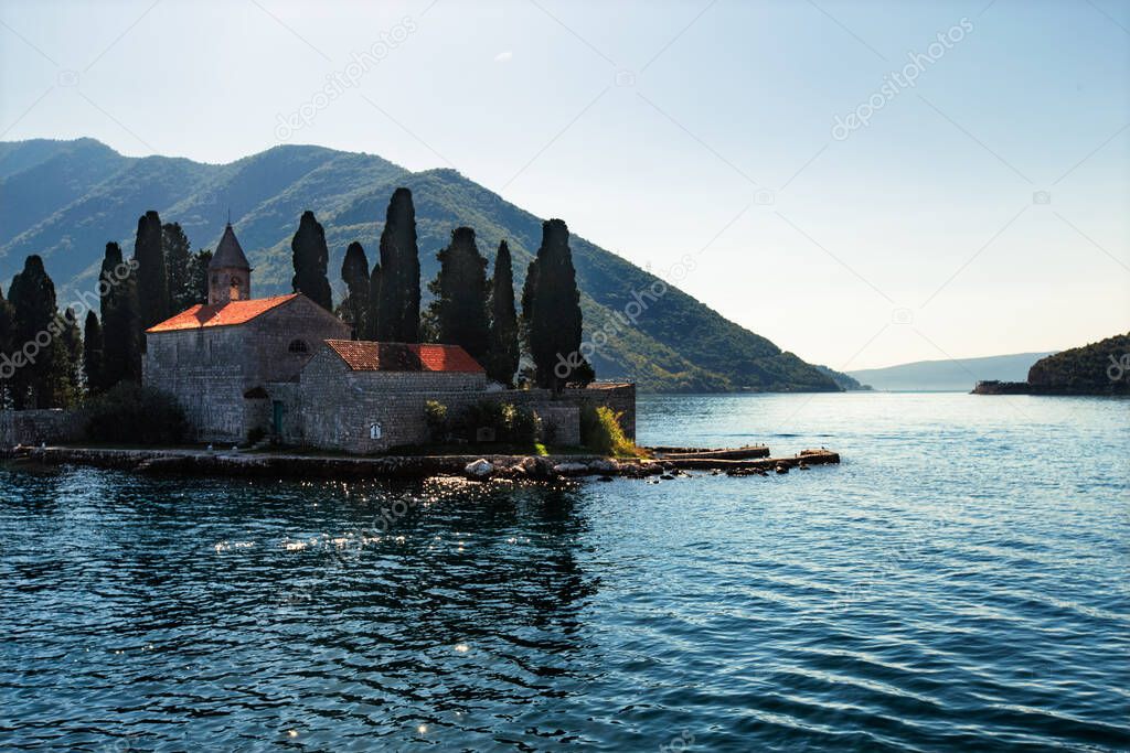 Catholic red roof medieval monastery, bell tower, masonry wall and cypress trees on an island in the Adriatic Sea.  Little island in blue water of Kotor bay among Montenegro mountains. Island panorama