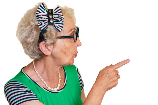 An old wrinkled hipster woman in glasses points her finger to the side with a surprised expression on her face. Isolate. Advertising space — Stock Photo, Image