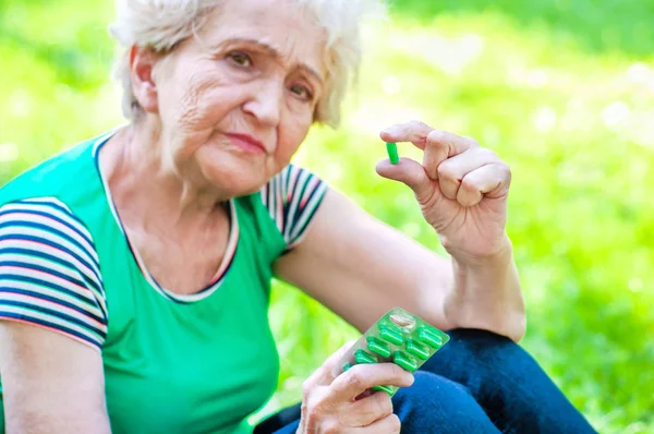 Грустная пожилая женщина держит в руках зеленые таблетки, сидя на лужайке. Здоровье женщин через 60 лет. Содействие здоровью в зрелом возрасте — стоковое фото