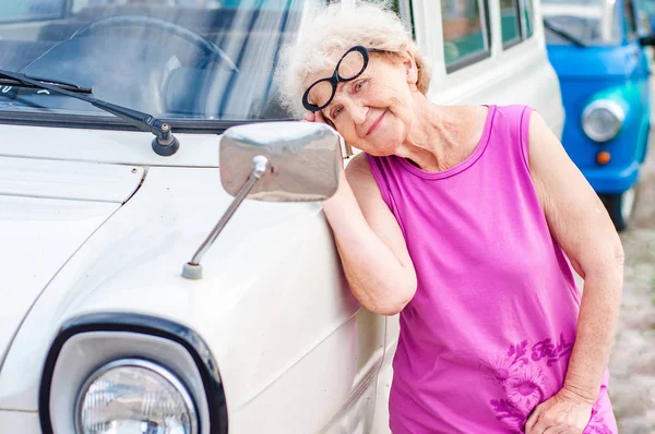 Модная привлекательная женщина в очках старше 60 лет смотрит на себя в зеркало ретро-машины. Летние каникулы и путешествия пожилых людей — стоковое фото