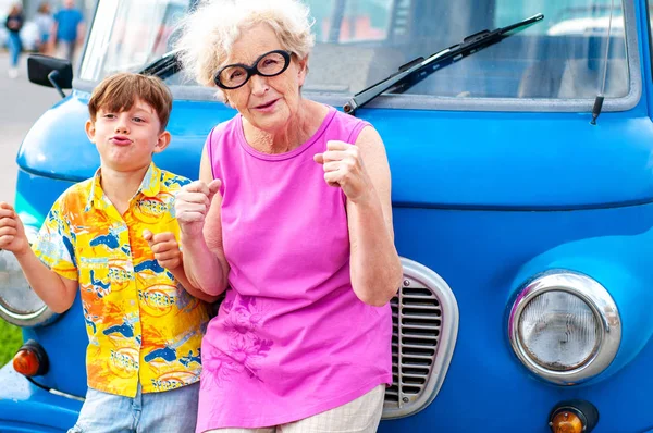 Modische, gut gelaunte Großmütter mit ihrem Großvater albern um ein Retro-Auto herum, das das Autofahren imitiert. Sommer Familienurlaub und Reisen — Stockfoto