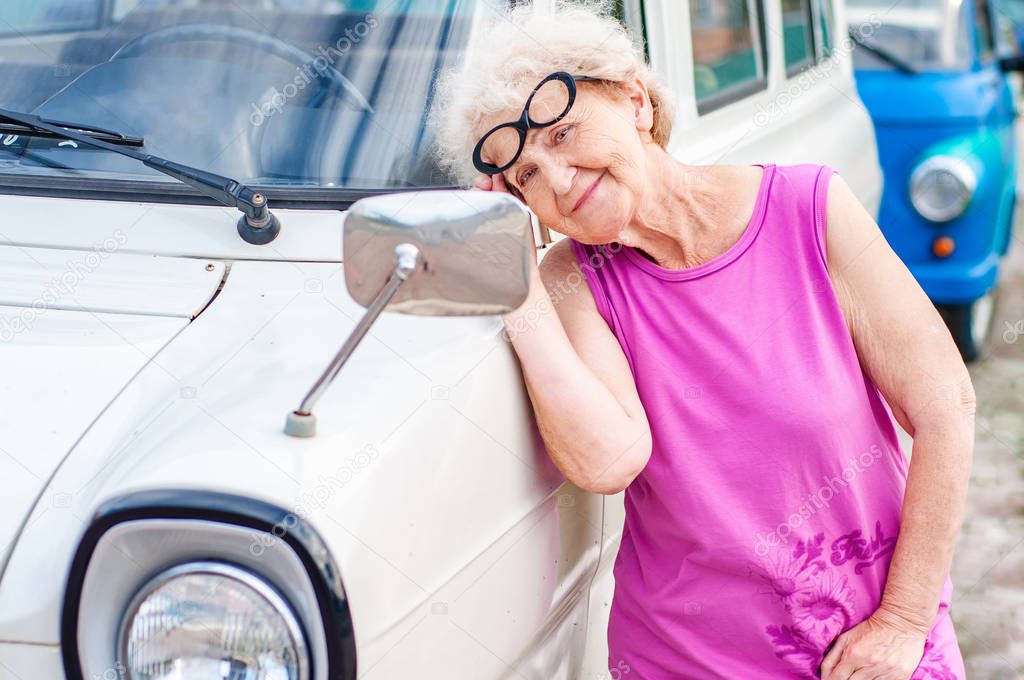 Fashionable attractive woman in glasses over 60 is looking at herself in the mirror of a retro car. Summer vacation and travel of older people