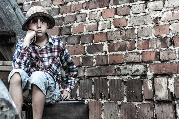 Arme trieste jongen zit op een koffer tegen een bakstenen muur. Dakloze eenzame kind — Stockfoto