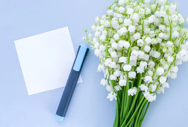 A bouquet of lily of the valley on a blue background, an empty sheet of paper and a marker. Top view, empty space for text.