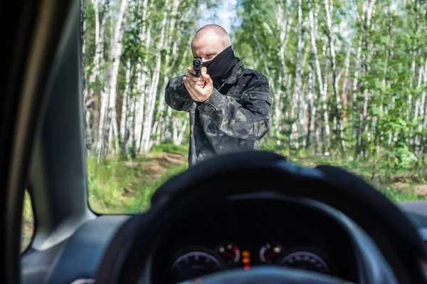 man in camouflage with a pistol aims at the driver of a car on the road in the forest