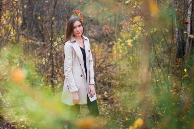 Gün batımında sonbahar ormanında yağmurluk giyen güzel, narin bir kız. Ilık ışık. Sarı ve kırmızı yapraklar