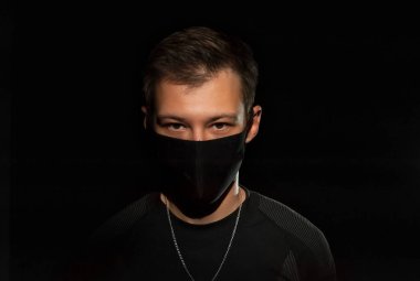 Siyah elbiseli ve siyah arka planda siyah tıbbi maskeli adam kameraya bakar. Yakın plan.