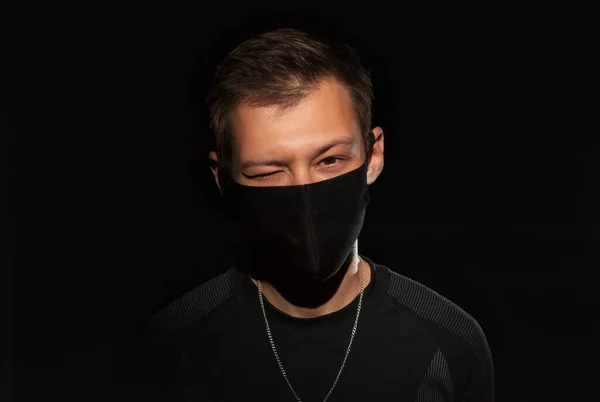 The guy in black clothes and a black medical mask on a black background winks and looks at the camera. Close-up