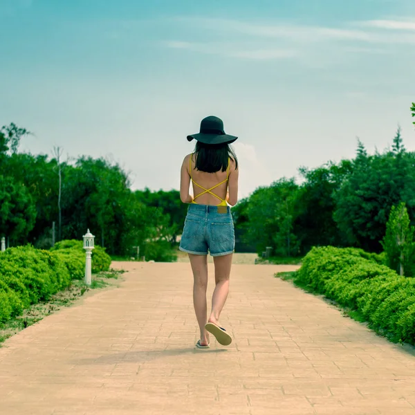 A vista da parte de trás da menina no corpo amarelo, calções jeans e chapéu, está na estrada no verde — Fotografia de Stock