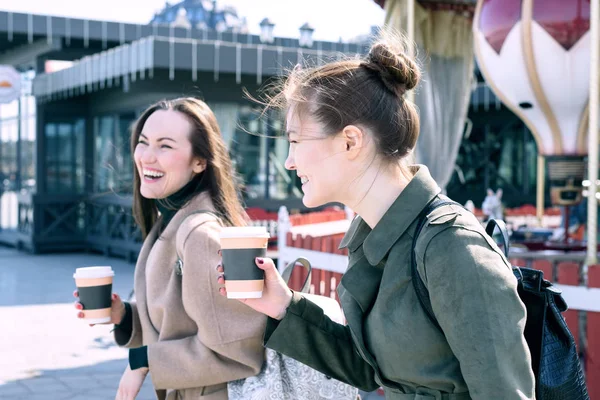 Счастливые две молодые женщины улыбаются тяжело и смеются ходить по улицам города в солнечный день, держа чашки кофе в руке — стоковое фото