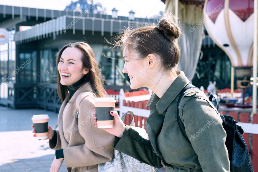Happy two young women are smiling heavily and laughing walking the streets of the city on a Sunny day, holding cups of coffee in his hand