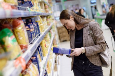 Smiling business woman in a jacket in a supermarket chooses a package of pasta. clipart