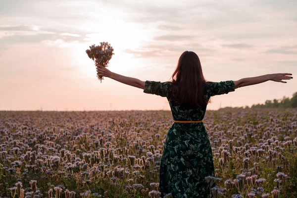 Freedom. Woman with open arms and a bouquet of flowers at sunset on flower meadow