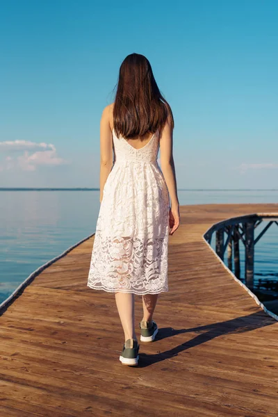 Vertical portrait of brunette in white sundress from the back is walk on wooden pier