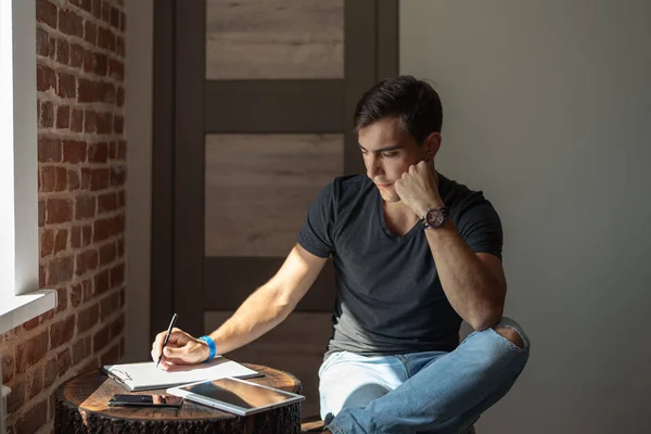 Thoughtful man writing a notebook next to tablet, sitting at table in room, work at home, planning, accounts, startup