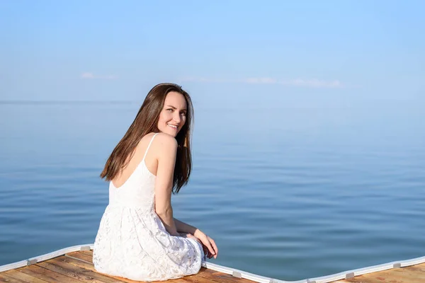 Brunette sitting on pier in white sundress, smiling, looking at camera in half turn on background of sea, rear view, copy space