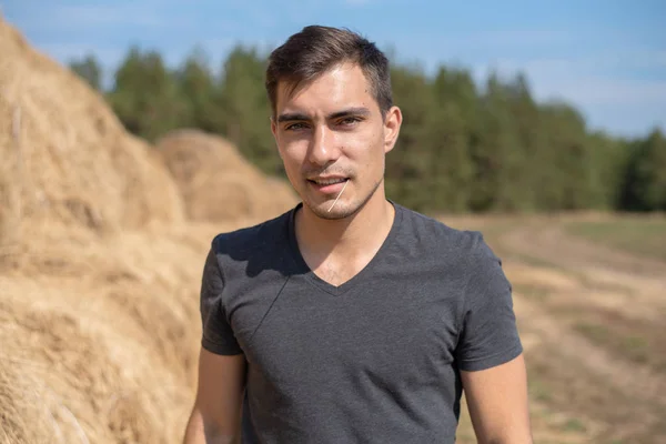 Frontal authentic portrait of young man in gray t-shirt on the background of field with haystacks