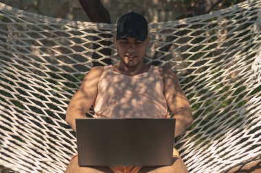 Genç adam bir kap, oturan bir hamakta bir dizüstü bilgisayar, serbest, 5 g bağlantı, çalışma tatil yazlık giysiler