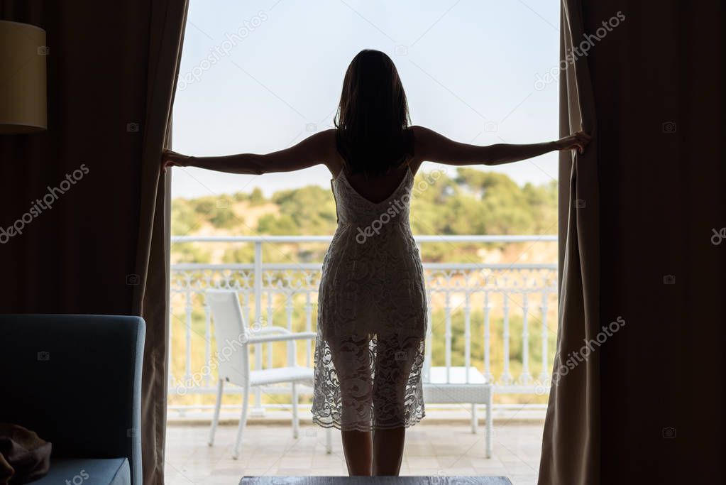 Young slender woman in a white dress opens the curtains of the panoramic window in the room. Meeting a new day, enjoying the morning on vacation.