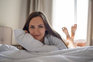 Frontal beyaz çarşaf ve yastık ile yatakta esmer kız portresi.