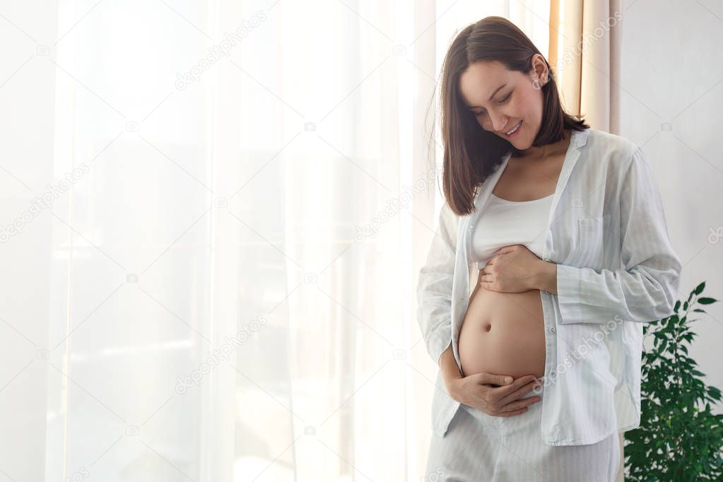 One beautiful pregnant girl standing at the window and looking with a smile on her belly. Happy motherhood concept. copy space