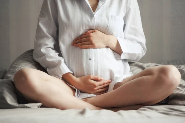閉じる - 座っているベッドの上にパジャマ姿で妊婦腹の女性のアップ — ストック写真