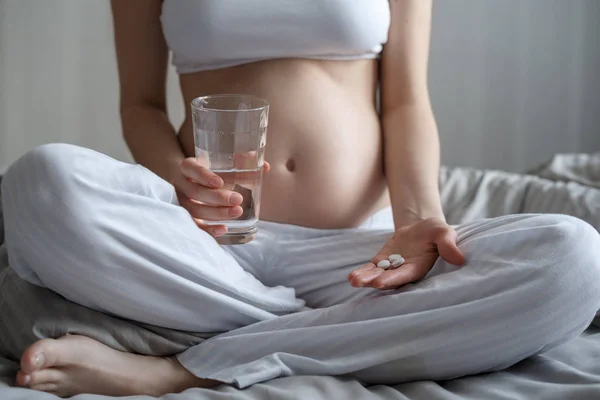 Закрыть беременную женщину с таблетками и стаканом воды, сидящей на кровати — стоковое фото