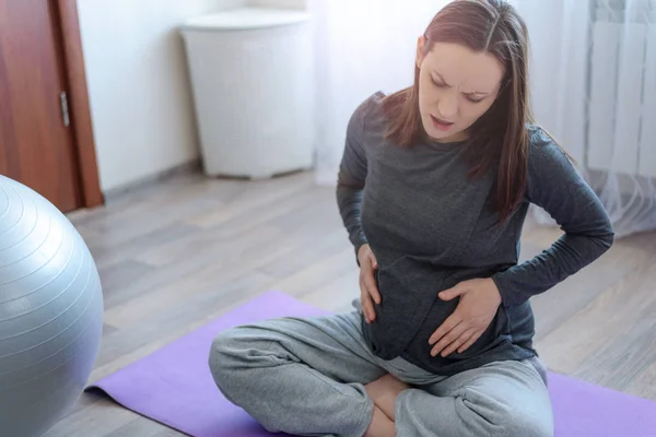 Pregnant woman experiencing pain sitting on the Mat with hands on her belly — Stock Photo, Image
