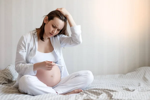 Donna incinta angosciata seduta sul letto tenendo un termometro e tenendo la testa. Il concetto di salute delle donne durante la gravidanza — Foto Stock