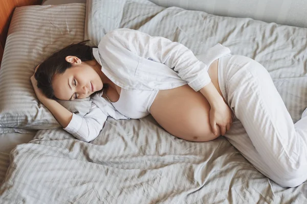 Giovane donna incinta sdraiata sul letto abbracciando la pancia, gravidanza sana — Foto Stock