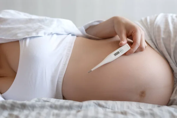 Close-up of the abdomen of a pregnant woman with an electronic thermometer in her hand. The concept of women\'s health during pregnancy