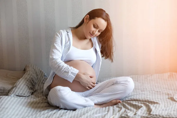 Feliz sorrindo jovem grávida sentada na cama e segurando a barriga. Saúde da mulher durante a gravidez — Fotografia de Stock