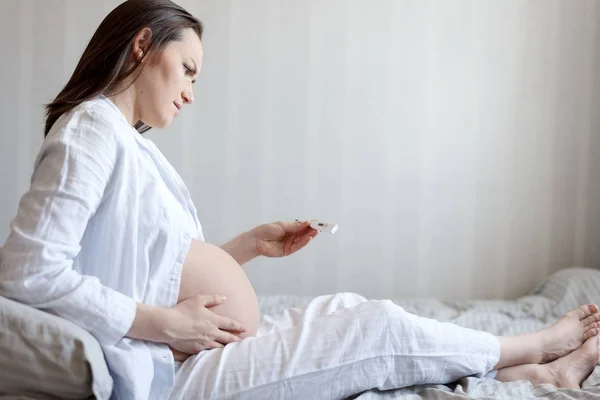 Vista laterale di una donna incinta a letto che tiene lo stomaco, guarda il termometro, freddo durante la gravidanza — Foto Stock