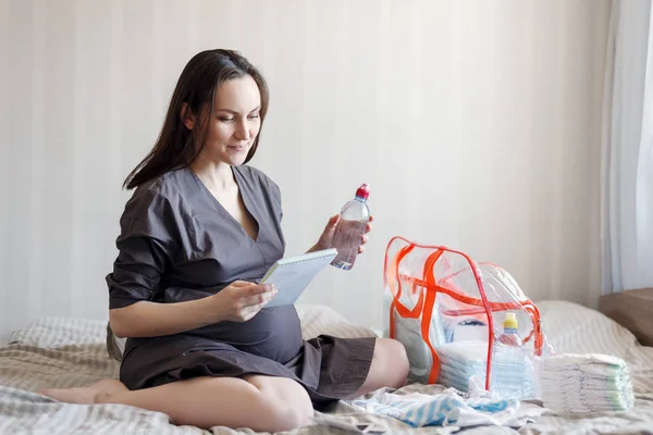 Mujer embarazada sonriente sentada en la cama con una lista en las manos, recoge cosas en la bolsa para el hospital — Foto de Stock