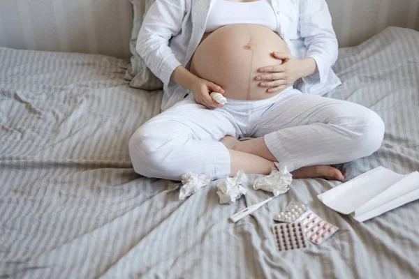 Primo piano di una donna incinta seduta sul letto, fredda, con spray nasale, pillole, fazzoletti di carta e un termometro. copia spazio — Foto Stock