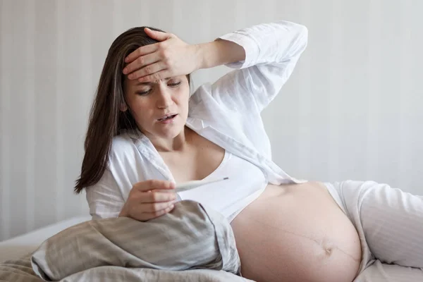 Ritratto frontale di una donna incinta sdraiata a letto con un termometro, tenendo la testa, la temperatura, il freddo, l'influenza durante la gravidanza — Foto Stock